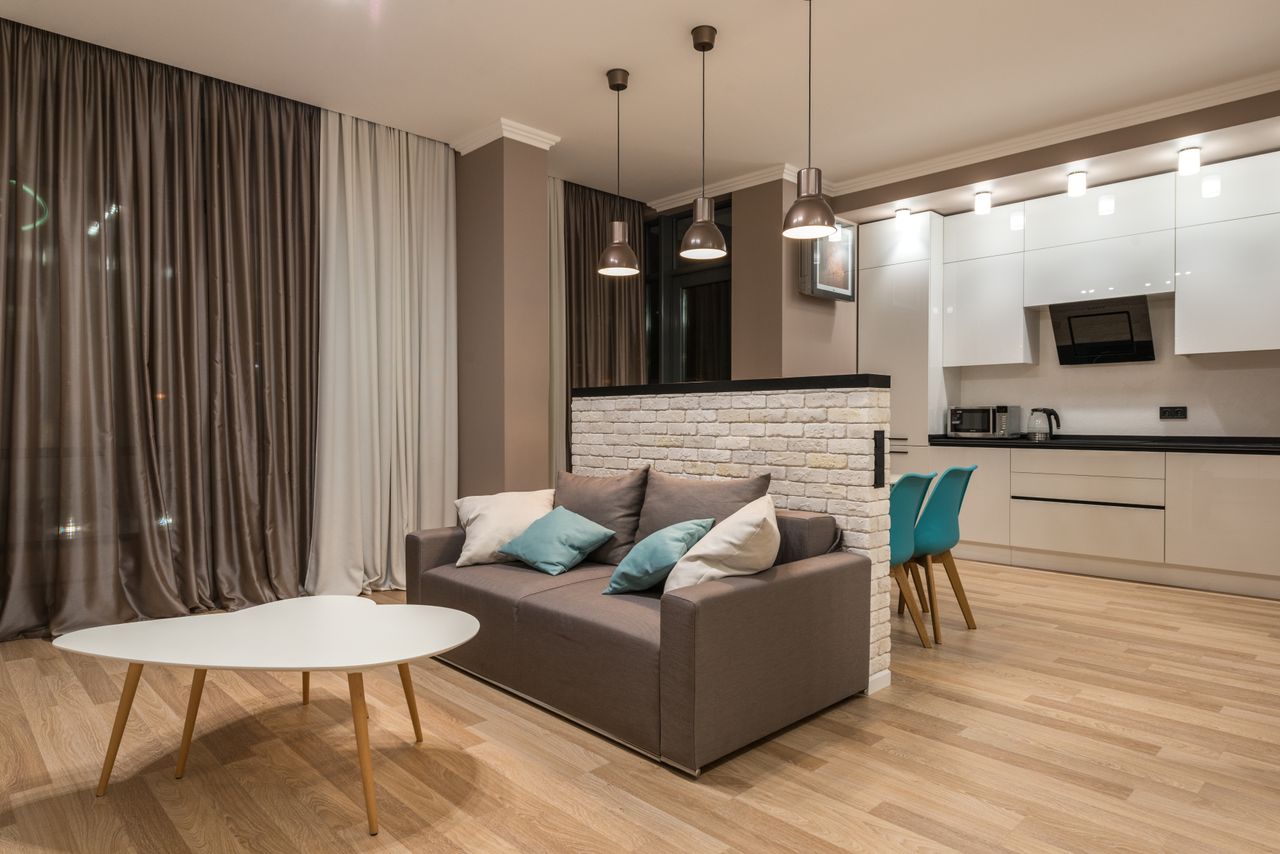 Interior of design of contemporary spacious living room with gray sofa and white coffee table and hanging lamps and joined furnished kitchen
